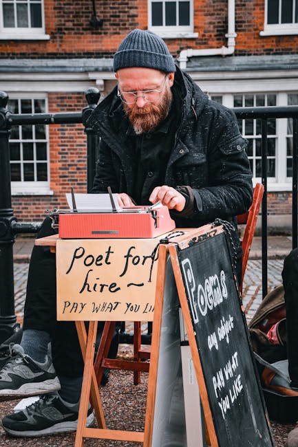 A man with a red beard wearing a dark knit beanie and black jacket is sitting outdoors at a small wooden table, writing on a pad of paper with a pen. The scene appears to be on a brick-paved area near a residential building with white-framed windows and black railings. In front of him, there is a cardboard sign that reads 'Poet for hire - Pay what you like,' indicating he is offering poetry services. Another blackboard sign with partial text is positioned nearby. The environment is overcast, with diffused natural light, and the focus is on the man engaged in his writing activity, suggesting a casual, outdoor street performance or promotional setup related to creative arts, not gardening or landscaping.