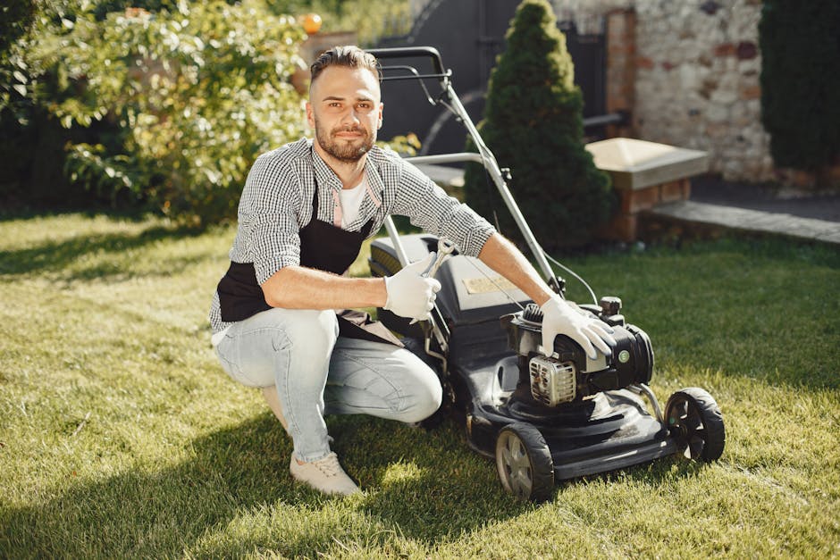 A man with neatly styled dark hair and a beard, dressed in a checkered shirt and light jeans, is crouching on a well-maintained green lawn while holding the handle of a black petrol-powered lawn mower with a visible engine. He is wearing white gloves and looking directly at the camera with a neutral expression. The garden setting features lush green grass, edged flower beds, and a brick pathway leading past trimmed hedges and a small conifer shrub. In the background, there is a yellow flowering shrub, a brick wall, and a stone feature, suggesting a tidy, landscaped outdoor space. The lighting indicates bright, sunny weather. This scene exemplifies garden maintenance work, highlighting lawn care and hedge trimming, and is associated with Gardens Ilford, a professional gardening service.
