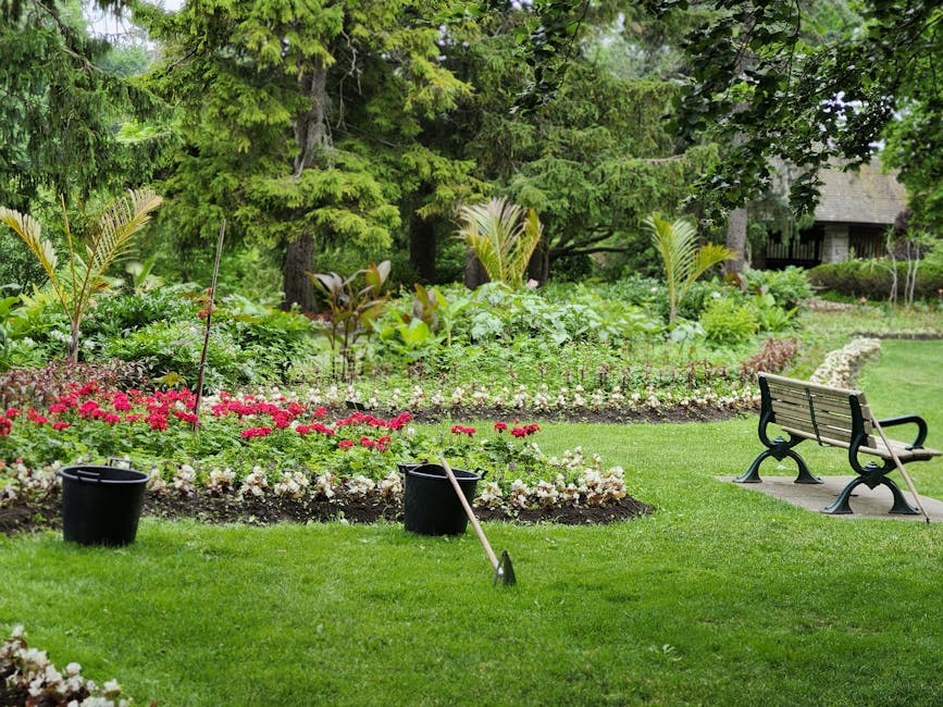 A well-maintained small garden scene featuring vibrant flower beds with pink and white blossoms, surrounded by lush green grass and leafy plants. The garden layout includes neatly bordered flowerbeds, and in the background, there are mature trees with dense foliage providing shade. A wooden bench with black metal armrests sits on a small paved area, offering a place for rest amid the garden scenery. Nearby, two black buckets and gardening tools such as a rake and a shovel are placed on the grass, indicating ongoing garden maintenance work. The environment appears fresh and tidy, with natural light illuminating the garden's variety of textures, including smooth soil patches, trimmed grass, and serrated leaves on the plants. The overall setting exudes a peaceful atmosphere suitable for landscaping work, hedge trimming, and lawn care, and it is associated with professional gardening services by Gardeners Ilford, evident from the scene's appearance and layout.